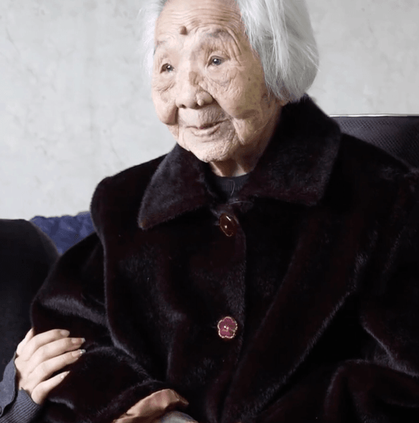 101-летняя долгожительница во время разговора с близкими Пожилая женщина в возрасте 101 года улыбается, сидя рядом с родственницей в домашней обстановке